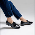 Black dress shoes worn with blue jeans on a light gray background, featuring the Caliber Shoes brand.