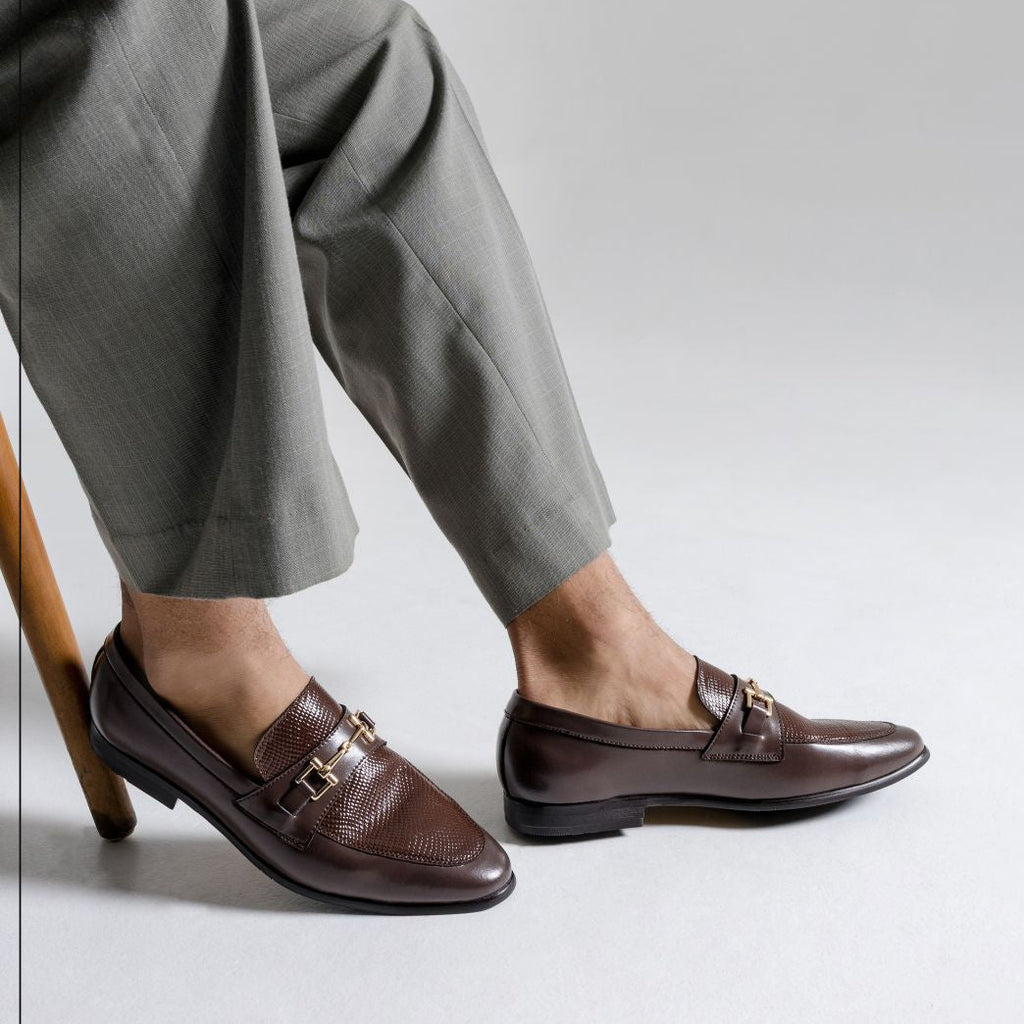 Brown loafers worn with gray pants on a light gray background, featuring Caliber Shoes logo.